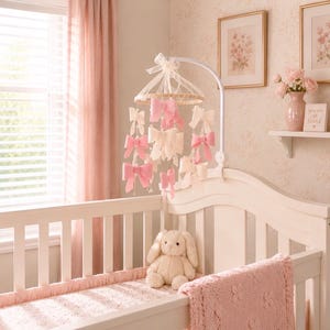 May include: A baby mobile featuring multiple hanging bows in shades of soft pink and cream, suspended from a circular frame with a white arm. The mobile is positioned above a white wooden crib containing a plush cream-colored stuffed bunny toy.