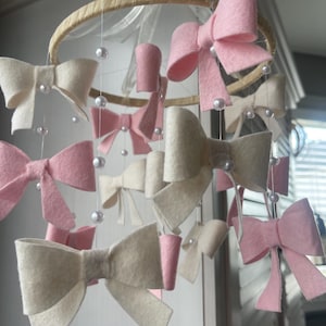 May include: A hanging mobile featuring multiple felt bows in shades of soft pink and cream, suspended from a circular wooden hoop by thin white strings. Small white faux pearl beads are interspersed along the strings between the bows.