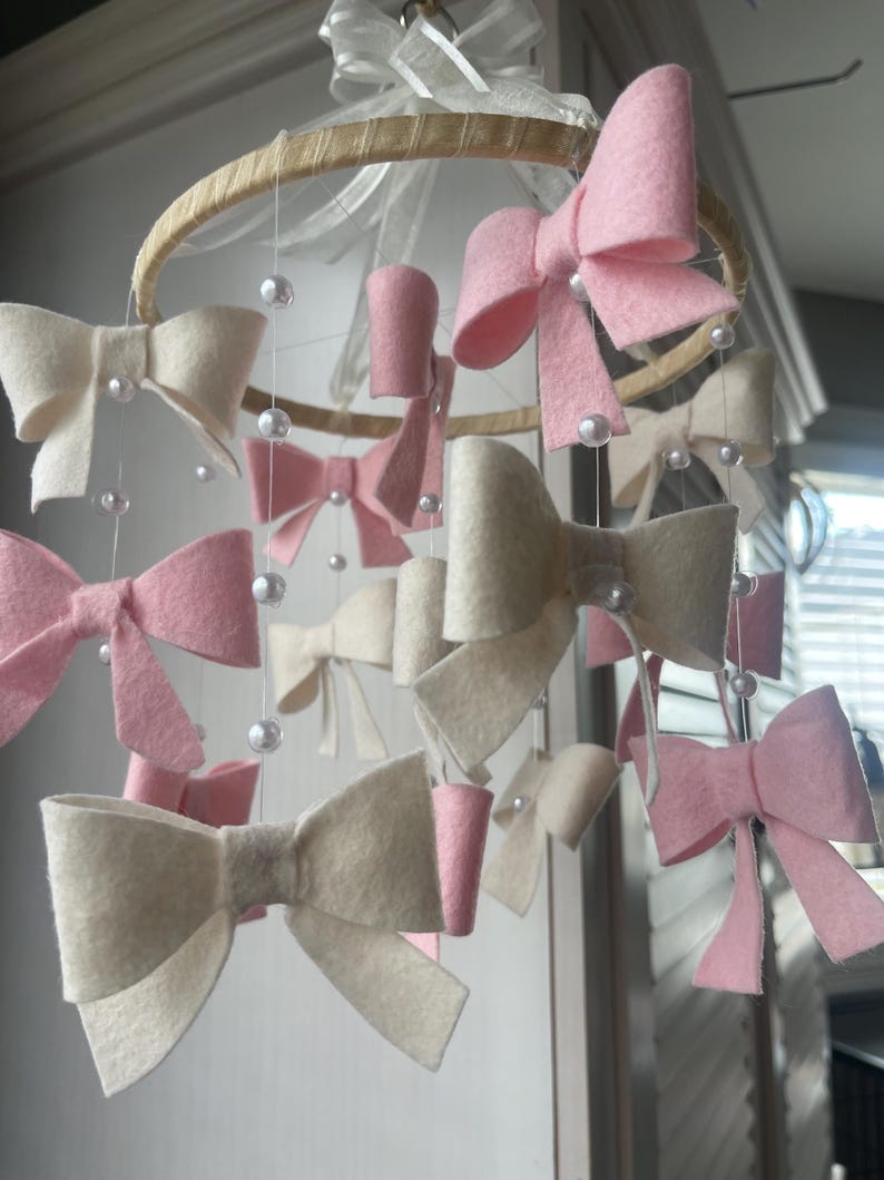 May include: A hanging mobile featuring multiple felt bows in shades of soft pink and cream, suspended from a circular wooden hoop by thin white strings. Small white faux pearl beads are interspersed along the strings between the bows.
