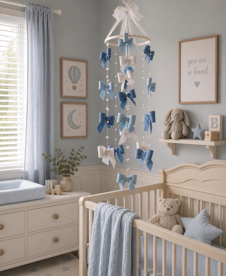 May include: A hanging nursery mobile featuring multiple fabric bows in shades of blue and white, suspended from a circular frame with white ribbons and strings of small white beads, creating a cascading decorative display above a crib.