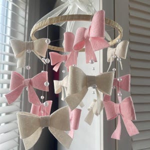 May include: A hanging nursery mobile featuring a circular wooden hoop wrapped in light fabric, adorned with multiple felt bows in shades of soft pink and cream. White faux pearl beads are strung on thin lines between the bows. Text reads Crib size.