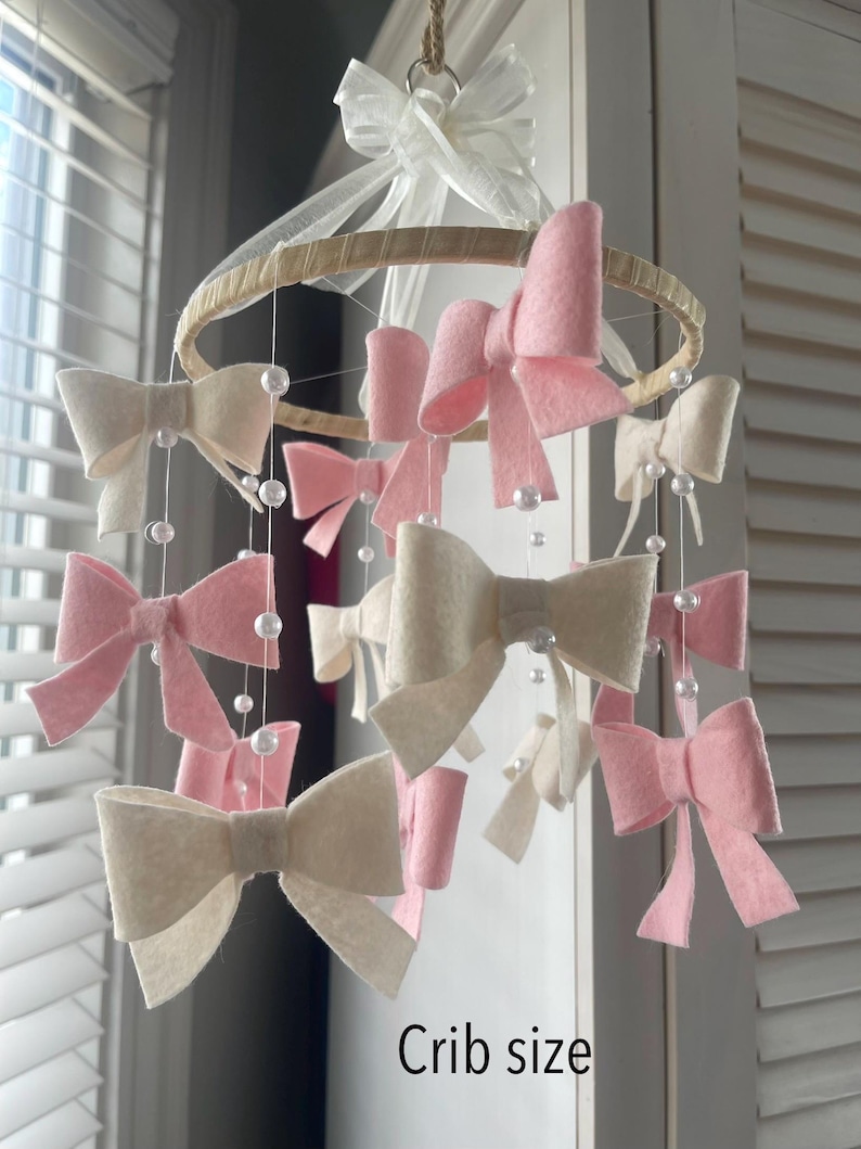 May include: A hanging nursery mobile featuring a circular wooden hoop wrapped in light fabric, adorned with multiple felt bows in shades of soft pink and cream. White faux pearl beads are strung on thin lines between the bows. Text reads Crib size.
