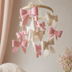 May include: A hanging mobile featuring a circular wooden hoop suspended by a sheer ribbon. Multiple felt bows in shades of soft pink and cream are attached to the hoop by thin strings accented with small white beads, creating a layered display.