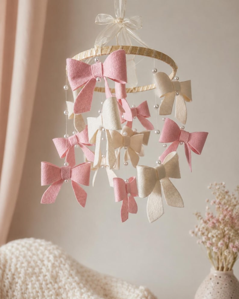 May include: A hanging mobile featuring a circular wooden hoop suspended by a sheer ribbon. Multiple felt bows in shades of soft pink and cream are attached to the hoop by thin strings accented with small white beads, creating a layered display.