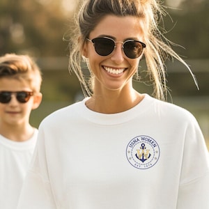 May include: A white sweatshirt with a navy blue circular logo that says "USNA WOMEN" and an anchor design. The sweatshirt is being worn by a person wearing sunglasses and blue jeans. A child is standing behind the person wearing a white t-shirt and sunglasses.
