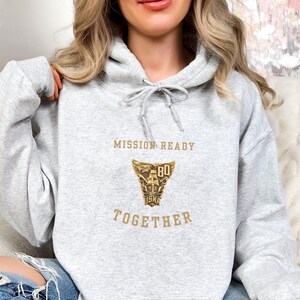May include: A pale grey hoodie with a drawstring and a front pocket. The hoodie displays the text "MISSION READY TOGETHER" and a gold emblem. The hoodie is made of a soft material, suitable for casual wear.