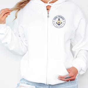 May include: White zip-up hoodie with a drawstring hood and a front pocket. The hoodie features a navy blue and gold emblem on the left chest that reads "USNA WOMEN EST. 1976" around an anchor and the letters "W" and "M".