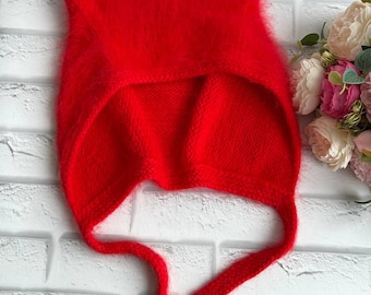 May include: A vibrant red knitted hat with ear flaps and long ties. The hat is made of a soft, fuzzy material and is designed to cover the head and ears. The hat is displayed against a white brick wall.