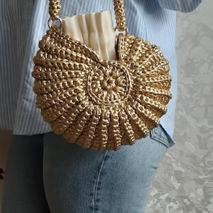 Könnte beinhalten: Eine goldfarbene, gehäkelte Handtasche mit kreisförmigem Design. Die Tasche hat einen passenden Griff und ein cremefarbenes Futter. Die Tasche wird von einer Person gehalten, die ein blau-weiß gestreiftes Hemd und eine blaue Jeans trägt.