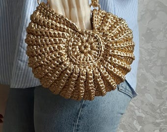 May include: A round, metallic gold crocheted handbag featuring a textured, ribbed shell-like pattern and a matching braided handle. The bag has a circular center detail and a light-colored fabric lining visible at the top opening of the purse.
