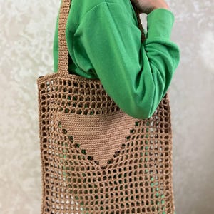May include: A brown, open-weave tote bag with a heart-shaped pocket. The bag has a rectangular shape with a woven strap. The bag is made of a light brown material and is designed for carrying items.