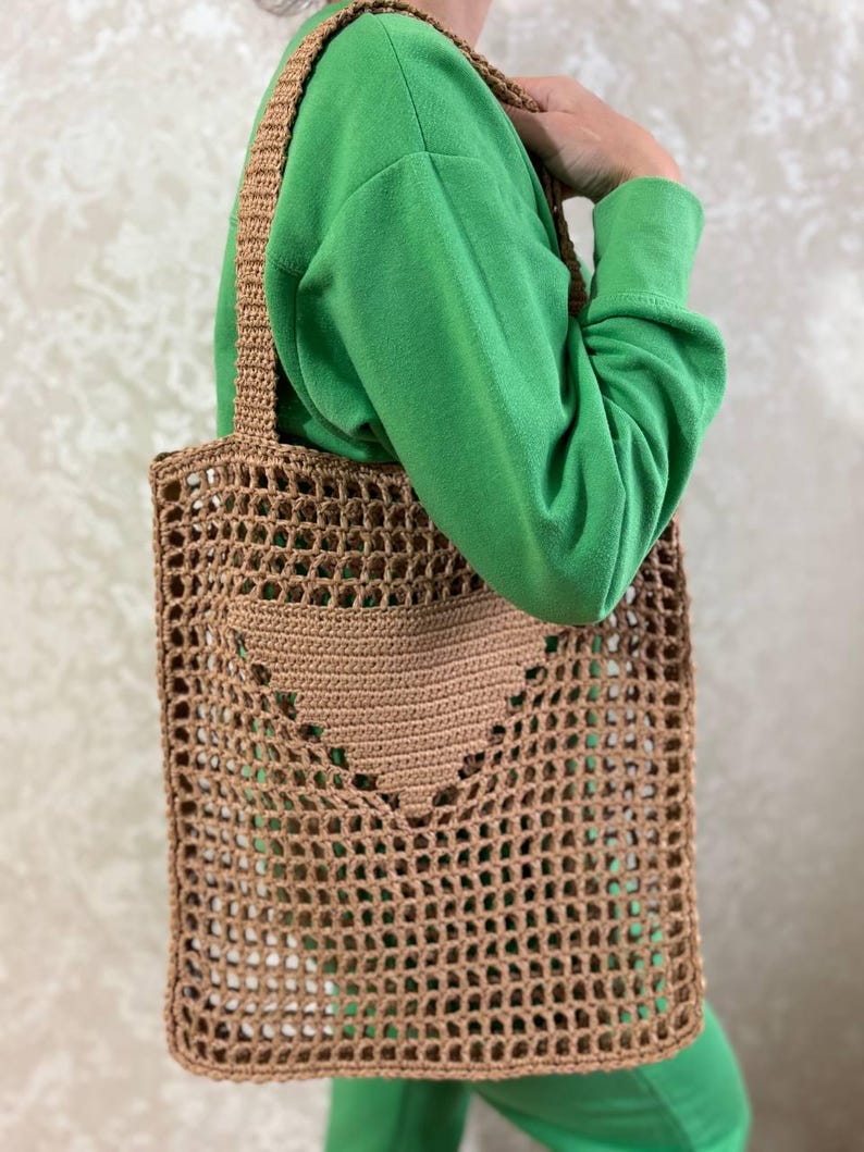 May include: A brown, open-weave tote bag with a heart-shaped pocket. The bag has a rectangular shape with a woven strap. The bag is made of a light brown material and is designed for carrying items.