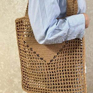 May include: A tan crochet tote bag with a large open weave design. The bag has a rectangular shape with a heart-shaped pattern in the center and two shoulder straps. The bag is made of a natural fiber material.
