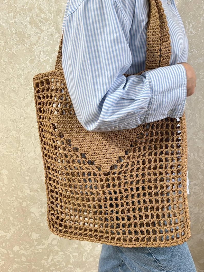 May include: A tan crochet tote bag with a large open weave design. The bag has a rectangular shape with a heart-shaped pattern in the center and two shoulder straps. The bag is made of a natural fiber material.