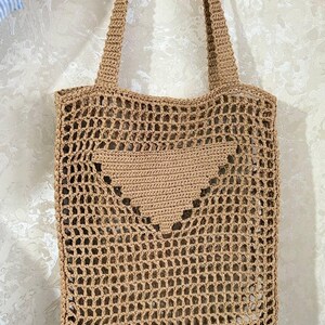 May include: A tan crochet tote bag with a heart-shaped design on the front. The bag has a rectangular shape with a mesh-like pattern and two shoulder straps. The bag is made of a natural fiber.