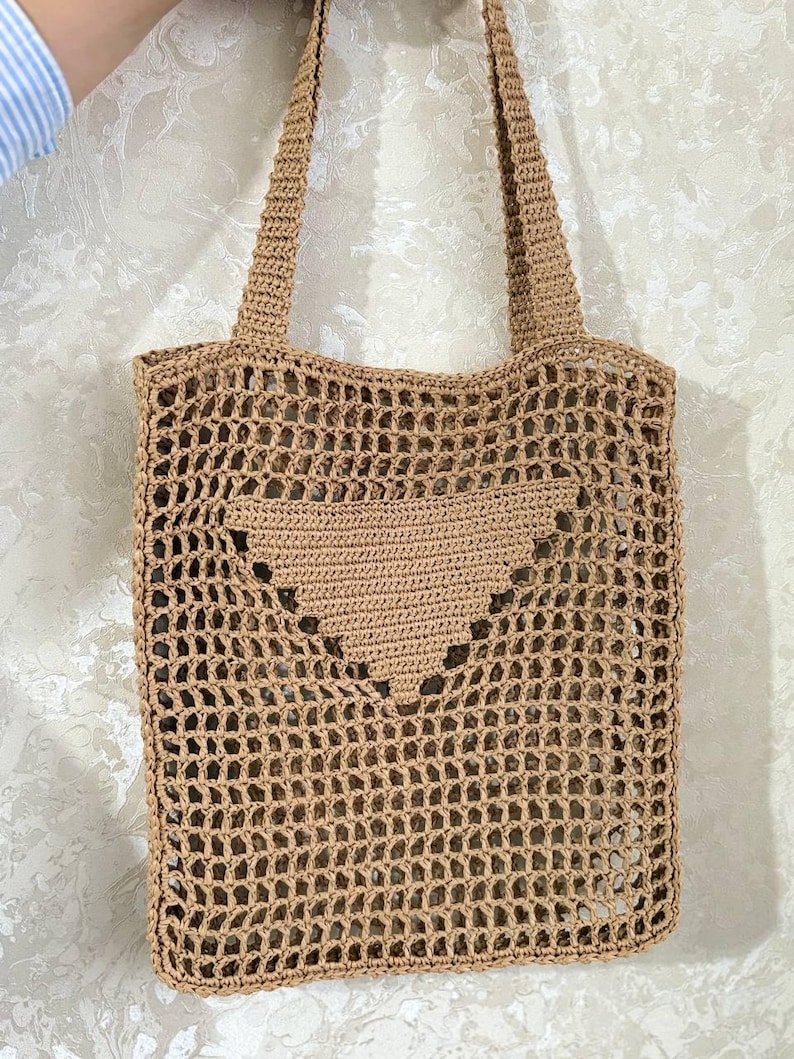 May include: A tan crochet tote bag with a heart-shaped design on the front. The bag has a rectangular shape with a mesh-like pattern and two shoulder straps. The bag is made of a natural fiber.