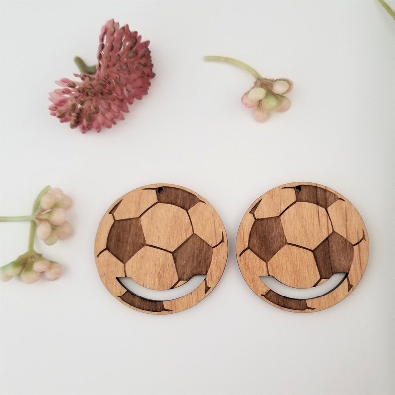 May include: A pair of wooden earrings shaped like soccer balls with a smile cut out of the bottom. The earrings are brown with a light wood grain.