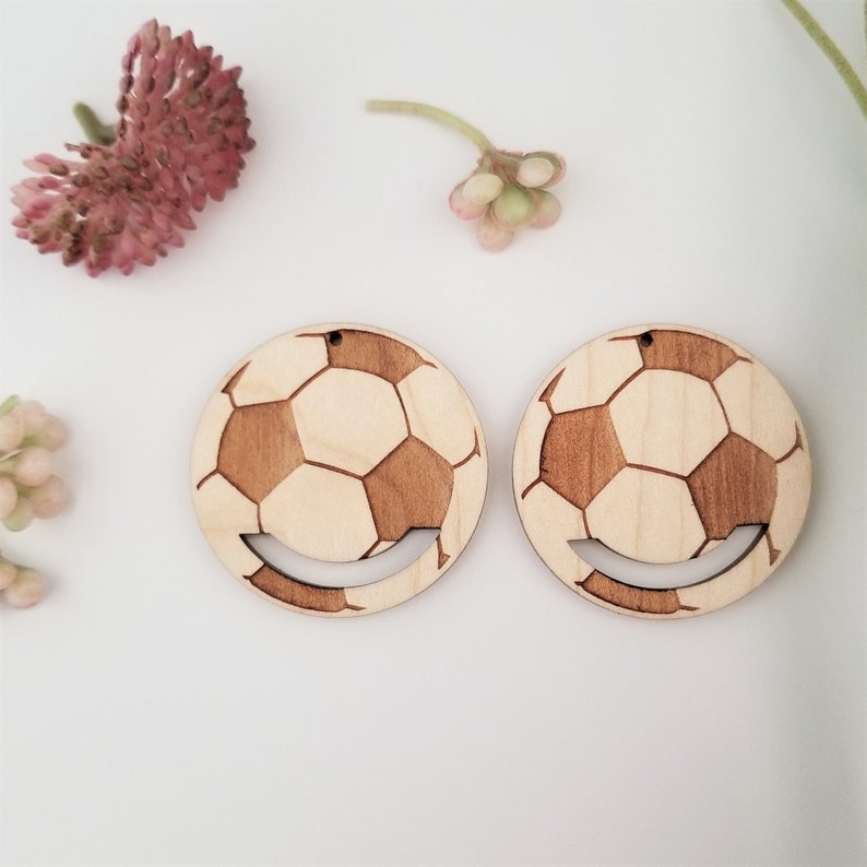 May include: A pair of wooden earrings shaped like a soccer ball with a smiling face cutout. The earrings are light brown with a natural wood grain.