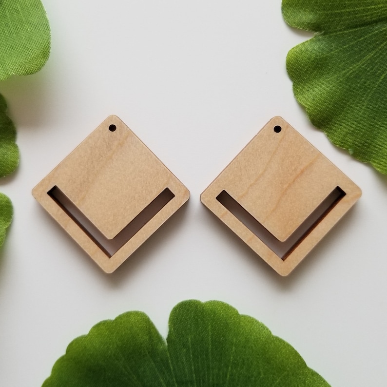 May include: A pair of wooden earrings with a diamond shape and a cut-out design. The earrings are a light brown colour and have a hole at the top for hanging.