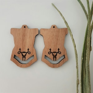 May include: A pair of wooden earrings shaped like a human torso with a stylized uterus and fallopian tubes design in the center.