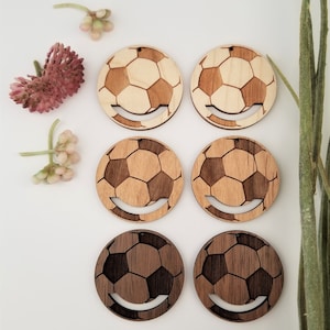 May include: Six wooden soccer ball earrings, three are light brown and three are dark brown. The earrings are round and have a smiley face cut out of the bottom.