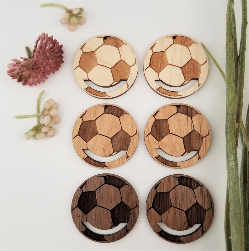 May include: Six wooden soccer ball earrings, three are light brown and three are dark brown. The earrings are round and have a smiley face cut out of the bottom.