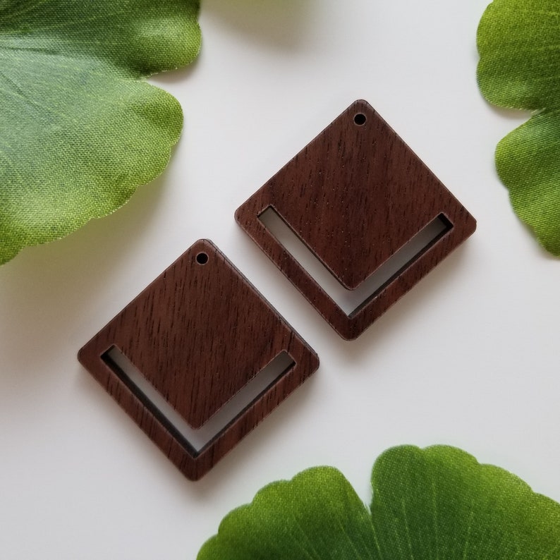 May include: Two dark brown wooden square earrings with a cut-out design. The earrings have a hole at the top for hanging.