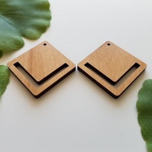 May include: Two wooden earrings in the shape of a square with a cut-out L shape. The earrings are a light brown colour with a dark brown border.