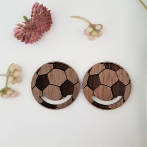 May include: A pair of wooden earrings shaped like soccer balls. The earrings are brown and have a wood grain texture. The soccer balls are cut out of the wood and have a white outline.