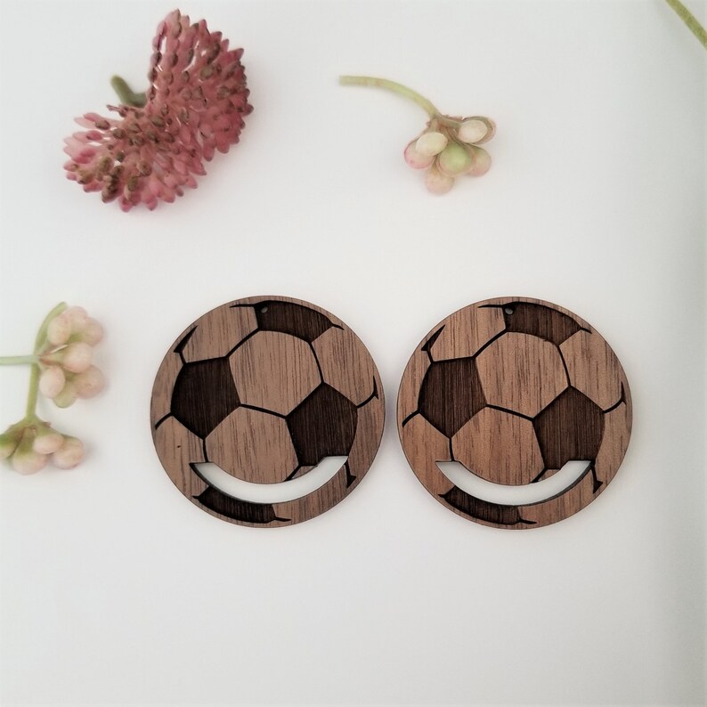 May include: A pair of wooden earrings shaped like soccer balls. The earrings are brown and have a wood grain texture. The soccer balls are cut out of the wood and have a white outline.