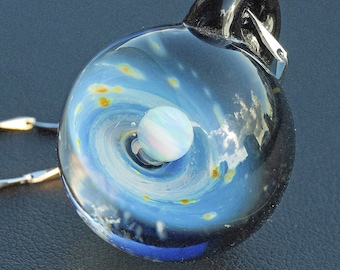 Puede incluir: Un colgante de vidrio con un diseño de galaxia en espiral y un pequeño planeta iridiscente en el centro. El colgante está unido a una cadena de plata.
