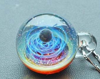 Puede incluir: Un colgante de bola de cristal con una galaxia azul y roja giratoria en el interior, una pequeña esfera negra que representa un planeta y una cadena plateada.
