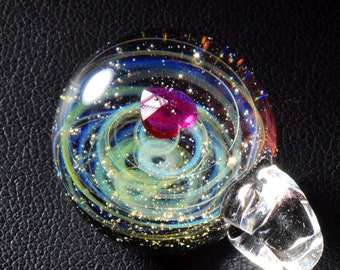 Puede incluir: Un colgante de vidrio con un diseño de galaxia en espiral y una piedra preciosa rosa en el centro.