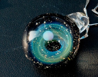 Puede incluir: Un collar con un colgante de vidrio con un fondo negro y un diseño de galaxia azul y verde en espiral. Una esfera blanca similar a una ópalo se encuentra en el centro de la galaxia.