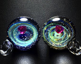 Puede incluir: Dos colgantes de vidrio con un diseño de galaxia en espiral y un corazón rosa en el centro. Los colgantes tienen un lazo plateado para colgar.