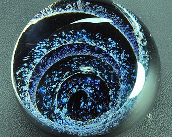 May include: A round glass paperweight with a swirling blue and black pattern. The pattern resembles a galaxy or a spiral.