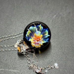 May include: A pendant necklace with a black glass orb, showcasing a vibrant floral design in blue, yellow, orange, and green. The pendant hangs from a fine silver chain. This unique necklace is a piece of jewellery.