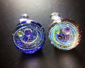 Puede incluir: Dos colgantes de vidrio con un diseño de galaxia. Los colgantes son azules y morados con remolinos dorados y pequeñas cuentas de vidrio verdes y moradas.