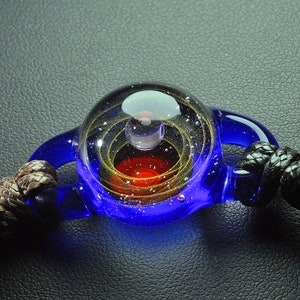 May include: A blue glass pendant with a red and gold swirl design inside. The pendant is attached to a black cord.