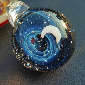 Könnte beinhalten: Ein Glasanhänger mit einem wirbelnden blauen und schwarzen Galaxie-Design und einem weißen Halbmond. Der Anhänger ist mit winzigen silbernen Glitzer gefüllt.