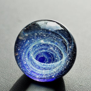 May include: A glass paperweight with a swirling blue and white galaxy design. The paperweight is clear and has a small, white letter "H" on the top.