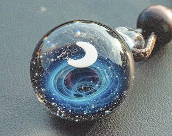 Puede incluir: Un colgante de cristal transparente con un diseño celestial. El colgante presenta una luna creciente blanca y un patrón de galaxia azul arremolinado con motas blancas. Cuelga de un cordón marrón.