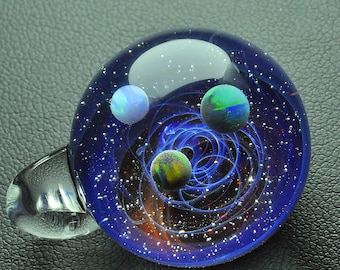 Puede incluir: Un colgante de vidrio con fondo azul y tres pequeñas esferas en su interior. Las esferas son iridiscentes y parecen ser planetas en una galaxia. El colgante tiene un lazo de vidrio transparente para colgar.