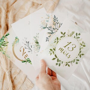 Peut inclure: Une main tient une pile de cartes blanches avec des illustrations botaniques. Les cartes présentent des motifs de feuilles vertes et dorées, avec du texte en or et vert. Une carte indique "13 mois" dans une couronne.