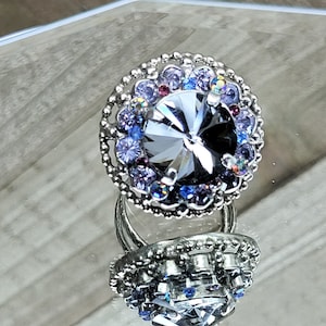 May include: A silver ring featuring a large, grey, faceted central stone. The ring is encircled by smaller, multi-coloured stones in shades of purple and blue. The ring is shown on a reflective surface.