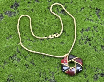 May include: A gold chain necklace with a geometric pendant. The pendant is made of black, red, and gold coloured materials and has a geometric design.