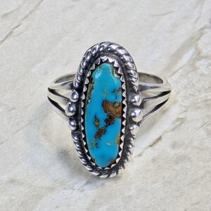 May include: A silver ring featuring an oval turquoise stone. The turquoise is a bright blue with brown markings. The ring has a twisted rope border and small silver beads. The band has a split shank design.