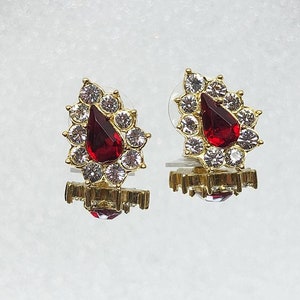 May include: A pair of gold earrings with a teardrop-shaped red stone surrounded by clear rhinestones. The earrings have a small gold base.