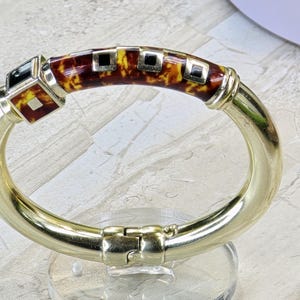 Puede incluir: Una pulsera de brazalete con bisagras doradas. La sección central presenta un patrón de carey con detalles cuadrados negros y amarillos. La pulsera se exhibe sobre un soporte transparente.