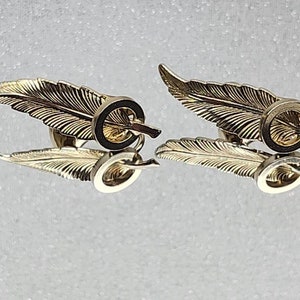 May include: A pair of gold-tone cufflinks featuring a feather design with a circular accent. The cufflinks are a classic and elegant accessory for any man's wardrobe.